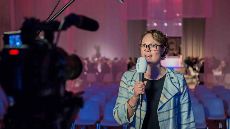 Vrouw met microfoon op podium, camera filmt haar, lege stoelen op achtergrond Vrouw spreekt met microfoon tijdens een evenement terwijl ze wordt gefilmd door een videocamera