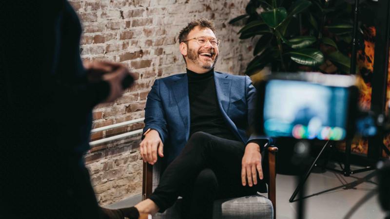 Man lachend in gesprek tijdens interviewopname, camera op de voorgrond Man zit ontspannen in een stoel en lacht tijdens een gefilmde interviewopname in een studio-omgeving