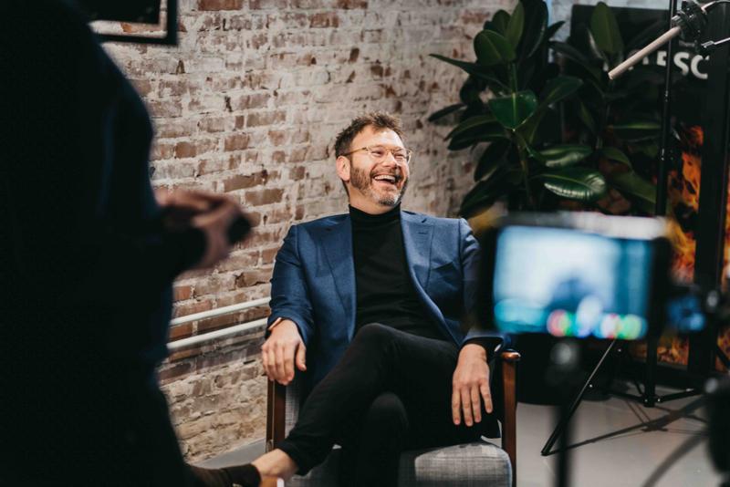 Man lacht tijdens een gefilmde interviewopname in een informele setting Man zit lachend in stoel, wordt gefilmd, camera zichtbaar op de voorgrond